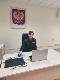 umundurowana uśmiechnięta policjantka siedzi za biurkiem przy komputerze, na ścianie godło Polski