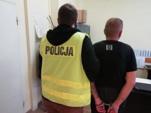 Mężczyzna z tatuażem na ręku, z kajdankami założonymi z tyłu i policjant w odblaskowej kamizelce z napisem Policja