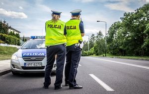 Policjant i policjantka w odblaskowych kamizelkach z napisem policja stoją na jezdni, w tle radiowóz