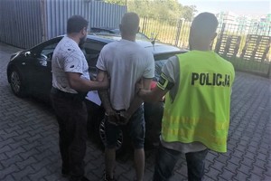 dwaj nieumundurowani policjanci prowadzą zatrzymanego mężczyznę do radiowozu