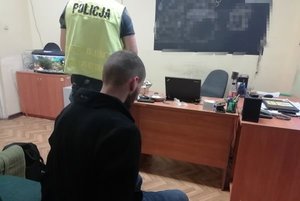 Siedzący mężczyzna z rękami z tyłu, dalej policjant przy biurku.