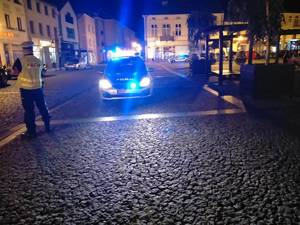 Zdjęcie wykonane o zmierzchu w Skierniewicach na miejskim Rynku. Na zdjęciu po lewej stronie stoi policjant ubrany w kamizelkę odblaskową, na środku zdjęcia widać policyjny radiowóz przodem, ma włączone światła i sygnały błyskowe.