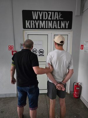 Policjant trzyma pod rękę mężczyznę z kajdankami na rękach, stoją przed drzwiami, nad drzwiami napis wydział kryminalny