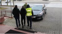 trzej policjanci doprowadzają do zaparkowanego przed komendą radiowozu, zatrzymanego będącego w kajdankach zespolonych