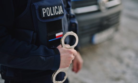 zdjęcie poglądowe, ręka umundurowanego policjanta trzymająca kajdanki, w tle radiowóz policyjny