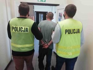 korytarz komendy, dwaj nieumundurowani policjanci w kamizelkach odblaskowych prowadzą zatrzymanego mężczyznę