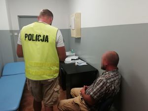 Mężczyzna siedzący na biurku, obok stojący policjant  w kamizelce z napisem policja