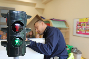 na pierwszym planie sygnalizator drogowy i światło zielone, w tle policjant rozmawiający z przedszkolakami