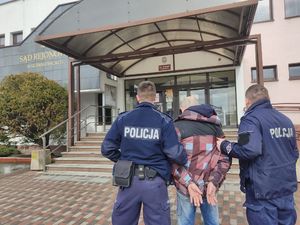 Meżczyzna z kajdankami na rękach,  dwóch policjantów przed budynkiem sądu