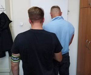 Mężczyzna stojący tyłem przed drzwiami, obok policjant po cywilnemu z opaską z napisem policja