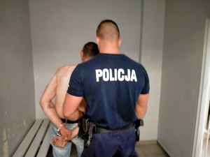 Policjant stoi tyłem, przed nim mężczyzna bez koszulki i z kajdankami na rękach.
