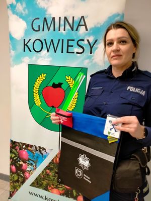 Policjantka trzymająca gadżety promocyjne na tle baneru z napisem Urząd Gminy Kowiesy