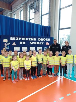 Dzieci oraz policjanci na hali sportowej na tle tablicy z napisem bezpieczna droga do szkoły