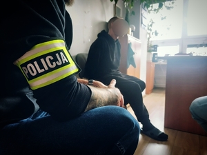 Policjant kryminalny, który ma na ręku opaskę policja, a w tle zatrzymany mężczyzna.