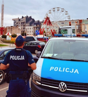 Policjant przy radiowozie a w tle karuzele, które są częścią imprezy masowej.