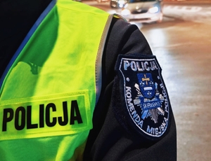 Napis policja na mundurze a w tle jadący pojazd.